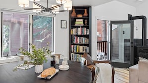 A dining area with a dark wooden table, white chairs, a chandelier, a bookshelf with books, and a view through large windows. - Outpost: Clark's Knoll 385 - Walk to Town Square (Jackson)