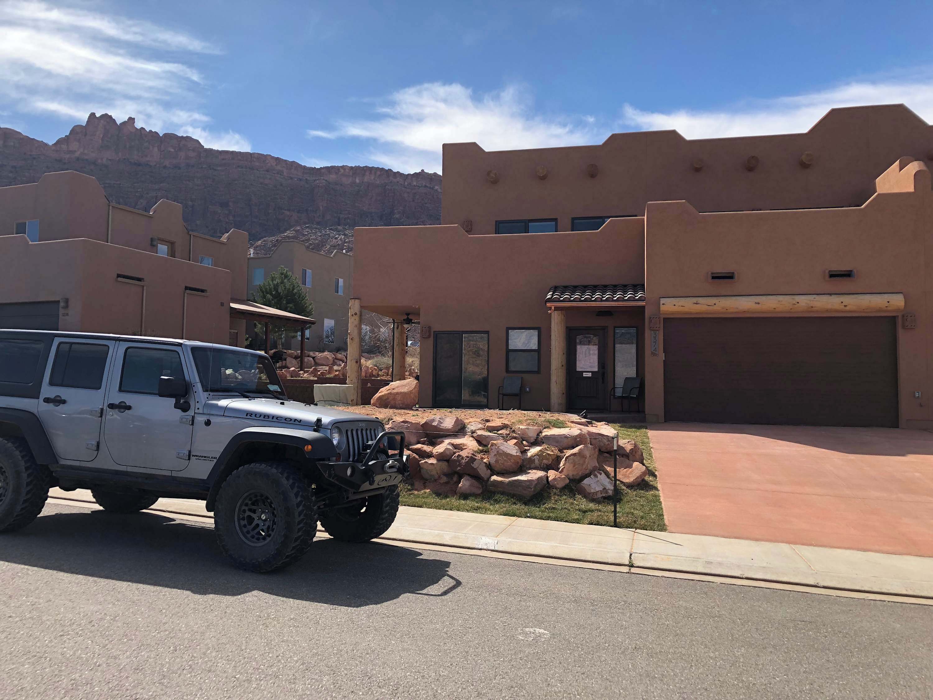 Un VUS blanc est garé devant un immeuble doté d’un garage, d’un espace patio avec des chaises et ayant une chaîne de montagnes en arrière-plan.