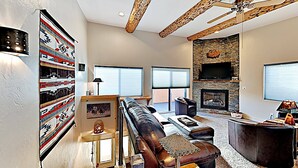 A modern living room with a sectional sofa, a fireplace, a television mounted on a stone wall, and a ceiling fan. - Moab Village Luxury Retreat Gorgeous views, PVT Hot Tub, 3 STE, 3.5 BTH, 1.5 KT (Moab)