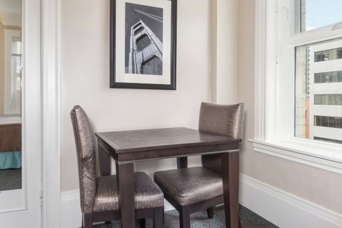 A dining area with a dark wooden table and matching chairs, a framed picture on the wall, and a window with a view of a building.