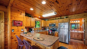 A wooden cabin interior with a kitchen featuring a central island, bar stools, and a welcome sign. - Resort setting w/ awesome Mtn views! Self checkin, close to Nat’l Park! Sleeps 6 (Pigeon Forge)