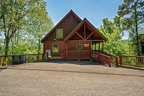 A wooden lodge with a covered porch, a fenced driveway, and a natural forest backdrop.