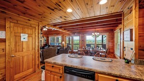 A wooden cabin interior with a kitchen area featuring a dishwasher, granite countertop, and a dining area with a table and chairs. - Resort setting w/ awesome Mtn views! Self checkin, close to Nat’l Park! Sleeps 6 (Pigeon Forge)