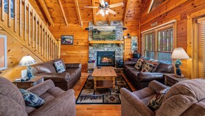 A log cabin interior with a fireplace, leather sofas, a wooden ceiling fan, and a stone fireplace surround. - Resort setting w/ awesome Mtn views! Self checkin, close to Nat’l Park! Sleeps 6 (Pigeon Forge)