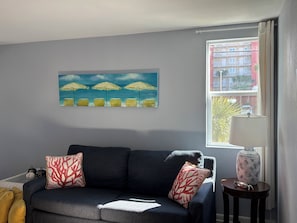 A living room with a dark blue sofa, two red and white patterned pillows, a painting of beach umbrellas, and a window with a view of buildings and a tree.