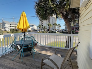 A wooden deck with a yellow umbrella, two chairs, and a view of a street with palm trees and buildings.