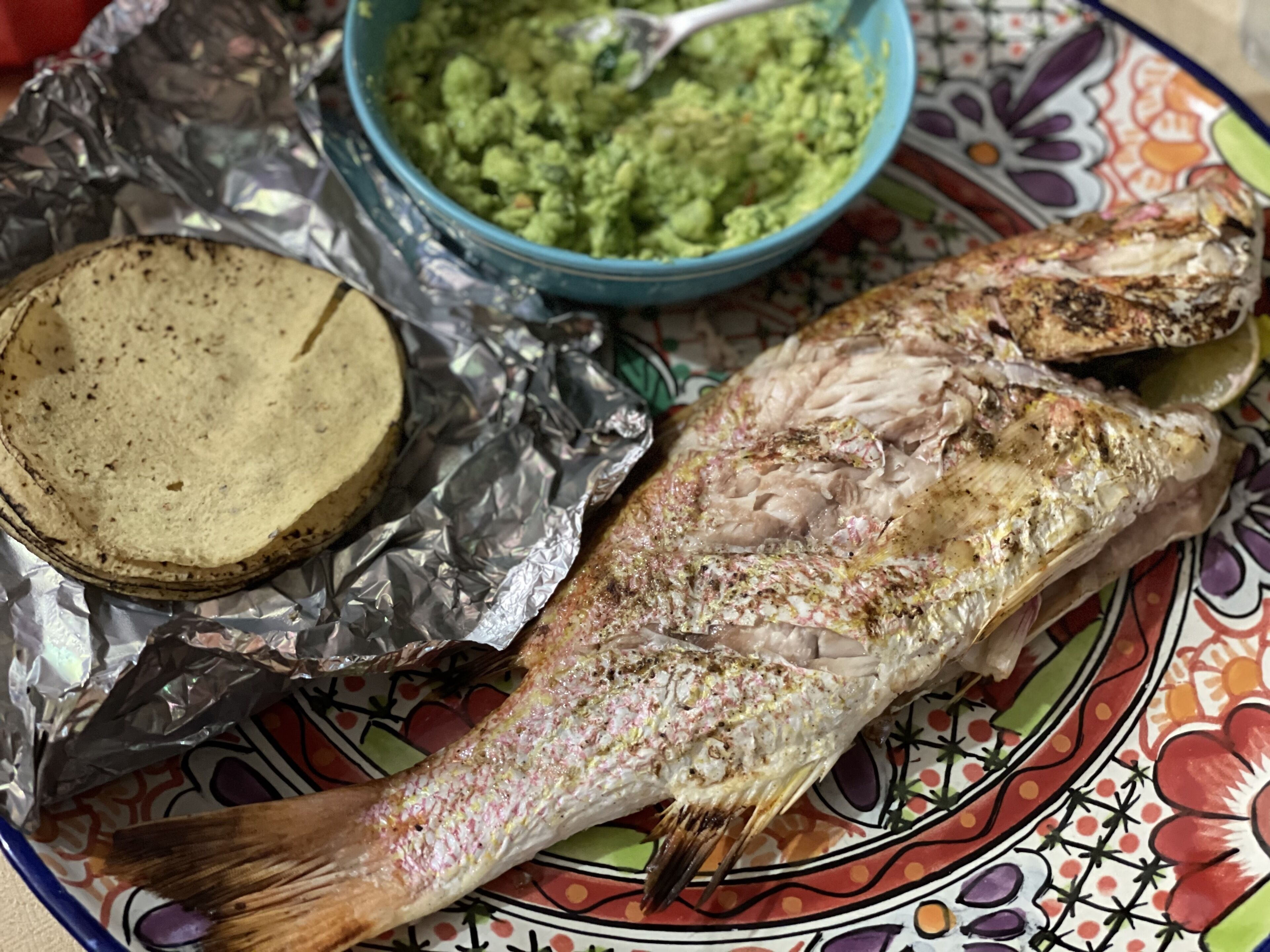 A whole fish on a colorful plate with foil wrapping and a bowl of guacamole.