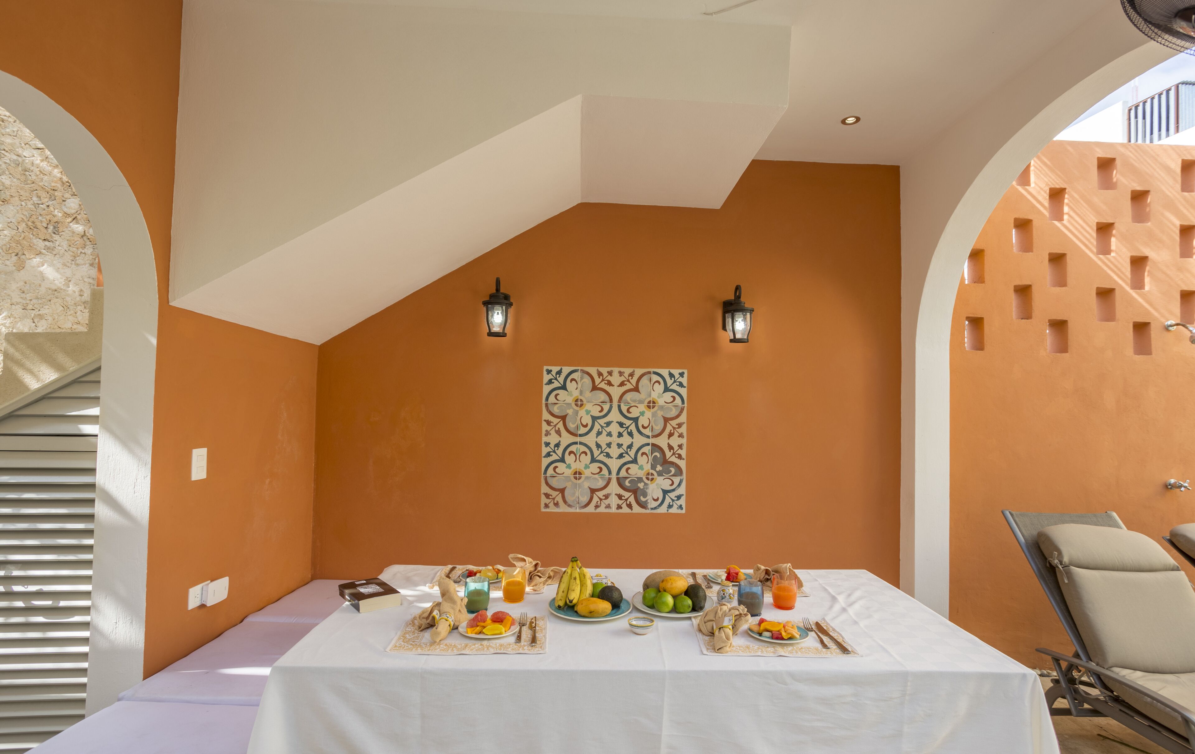 A dining area with a table set for breakfast, including fruits, a jug of juice, and a plate of eggs. There are two wall-mounted lanterns and a decorative tile above the table.