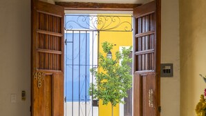 An open wooden door leading to a street with a yellow building and a tree.