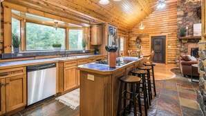 Een interieur van een blokhut met een keuken, voorzien van houten kastjes, een vaatwasser en een centraal kookeiland met krukken.