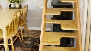 A wooden ladder with multiple steps leading to a white railing, a dining table with chairs, and a floral-patterned rug. - Layton's Loft (Manti)