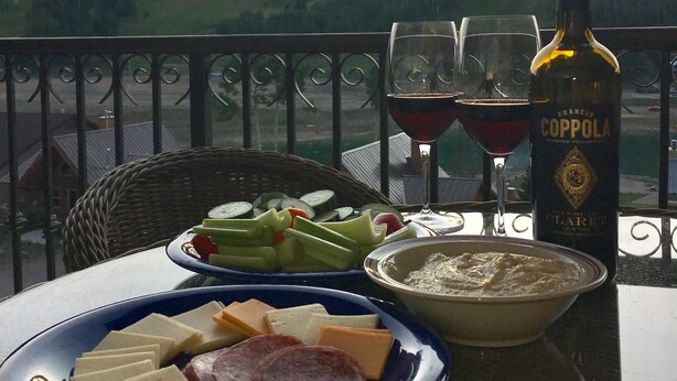 A table with a bottle of Franci Coppola wine, two glasses of red wine, a plate of cheese and cured meats, and a bowl of soup on a balcony with a scenic view of hills and a sunset. - Luxury Condo in Brian Head,  Beautiful Amenities,  Easy Walk/Ski To Slopes (Brian Head)