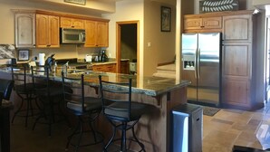 A kitchen with wooden cabinets, a stainless steel refrigerator, a microwave, and a coffee maker. There are four bar stools and a granite countertop. - Luxury Condo in Brian Head,  Beautiful Amenities,  Easy Walk/Ski To Slopes (Brian Head)