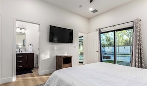 A bedroom with a large bed, a TV mounted on the wall, a bathroom with a mirror and sink, and a sliding glass door leading to a backyard.