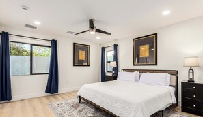 Une chambre à coucher comprenant un lit, une table de chevet, une lampe, un ventilateur de plafond et deux œuvres d’art encadrées accrochées au mur.