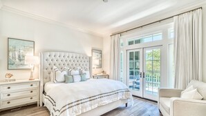 A bedroom with a tufted headboard, a large bed with white bedding, a nightstand with a lamp, a chair, and a view through a glass door to an outdoor area.