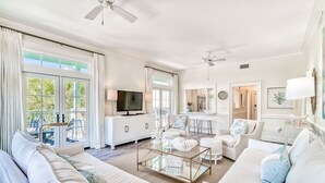 A spacious living room with a large TV, white sofas, a glass coffee table, and a view of the outdoors.