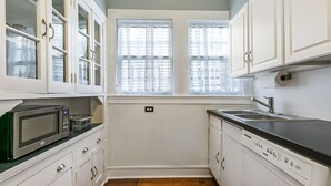 A compact kitchen with white cabinetry, a microwave, a sink, and two windows with blinds. - Riverbend Gem in Historic Carrollton Uptown  (New Orleans)