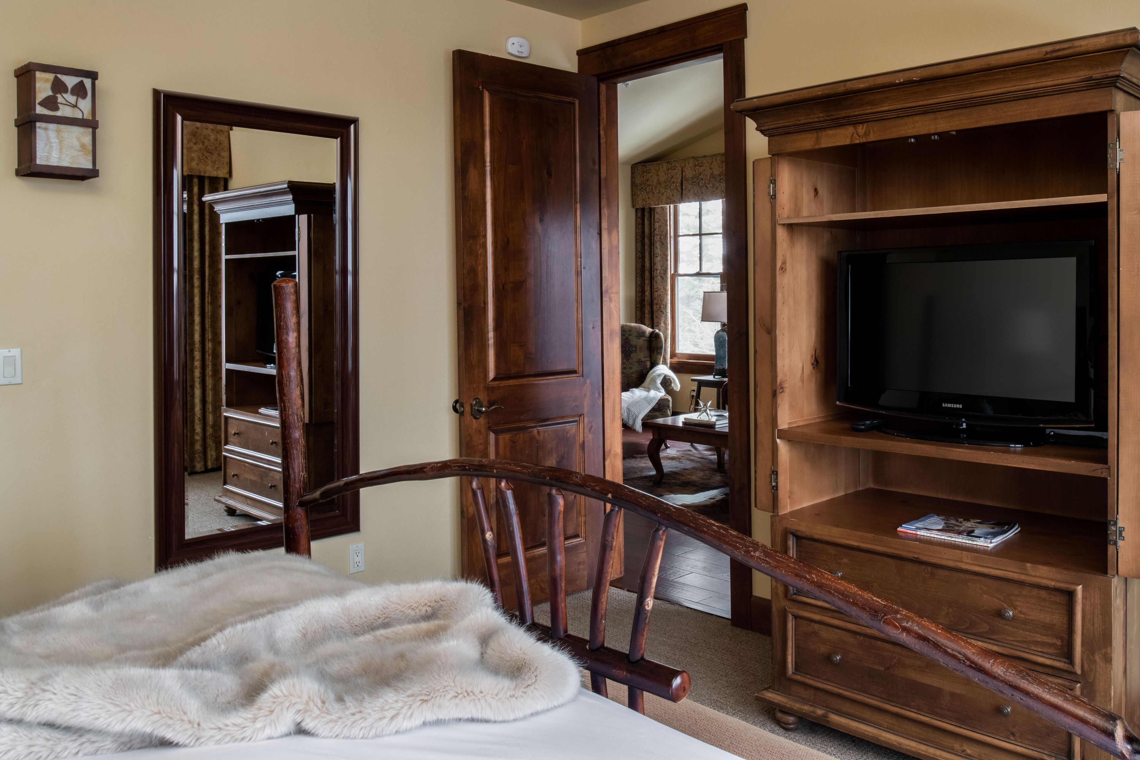 Une chambre avec une chaise en bois, un plaid en fourrure, un meuble TV en bois et un miroir qui reflète un placard et une chaise.