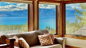 Habitación con ventana grande que ofrece vista a un lago y montañas. Hay un sofá con cojines estampados.