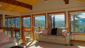 Una habitación amplia con techo alto, ventanas grandes que ofrecen vistas a una cadena montañosa boscosa, un sofá beige con cojines estampados y una pequeña mesa redonda.