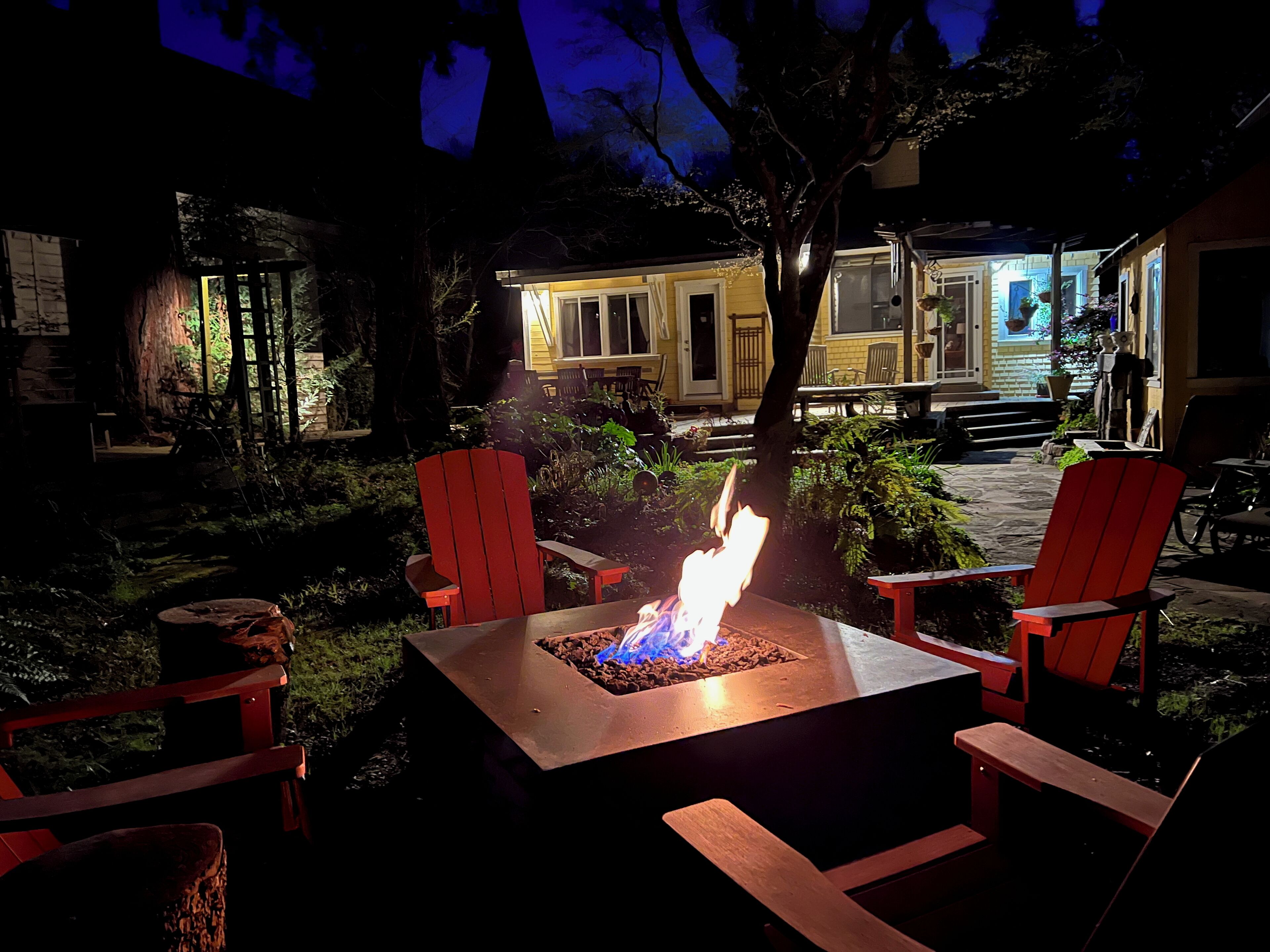 A fire pit with red chairs in a garden area, surrounded by trees and a building with a porch.