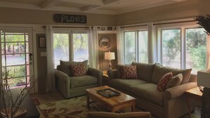 A cozy living room with two sofas, a coffee table, and a lamp. There are large windows with curtains, a door with a sign, and a wall clock. - The Kenwood House - A Place to Stay
Your Wine Country Oasis in Sonoma Valley (Kenwood)
