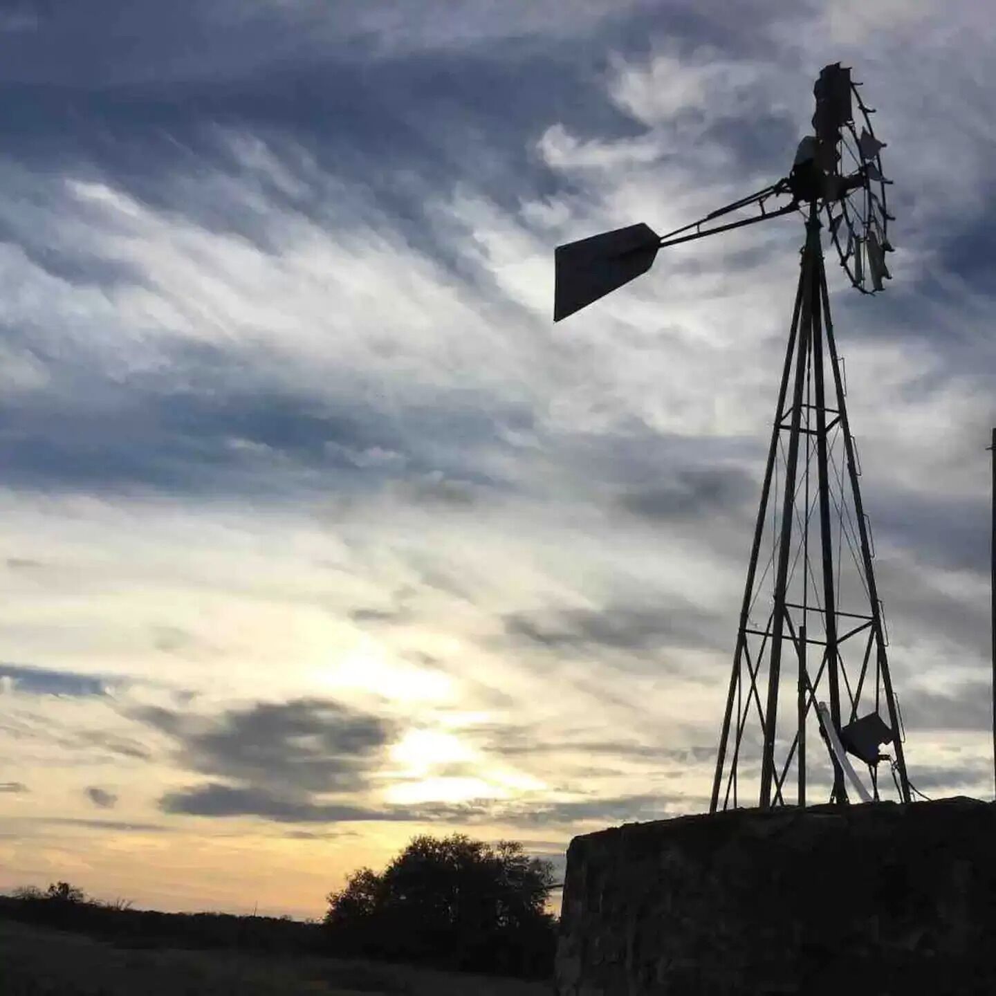 A windmill with a metal structure and a large fan at the top, set against a cloudy sky.