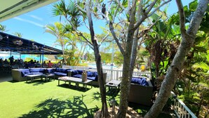 Un espace extérieur aménagé avec des canapés et des tables bleus, dotés de coussins, entouré d’arbres et avec une piscine en arrière-plan.