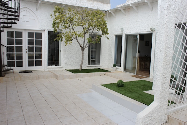 Un edificio blanco con un patio empedrado, un árbol pequeño y una puerta.