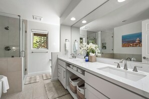 A modern bathroom with a large mirror, two sinks, a shower, and a window.