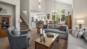 Een ruime woonkamer met een grijze bank, een houten salontafel met boeken, een grijze fauteuil en een trap naar een keuken.