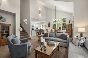A spacious living room with a grey sofa, a wooden coffee table with books, a grey armchair, and a staircase leading to a kitchen.