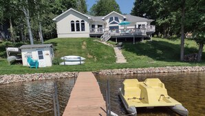Una casa con un muelle que se adentra en un lago, una pequeña embarcación amarrada a la derecha y una zona cubierta con sillas a la izquierda.