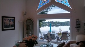 Una sala de estar acogedora con un amplio ventanal con vistas a un lago, un reloj y un jarrón con flores sobre una mesa.