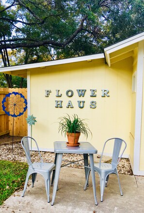Un edificio amarillo con la inscripción 'FLOWER HAUS' en la pared, dos sillas de metal, una mesa y una planta en maceta.