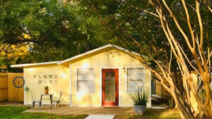 A yellow building with a red door, labeled "Flower Haus," featuring a white picket fence, a small table with chairs, and a potted plant.
