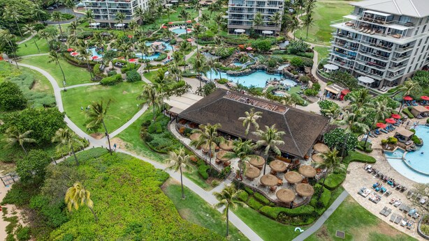 Eine Luftaufnahme zeigt ein Resort mit mehreren Schwimmbecken, Palmen und einem zentralen Gebäude mit einem Reetdach.