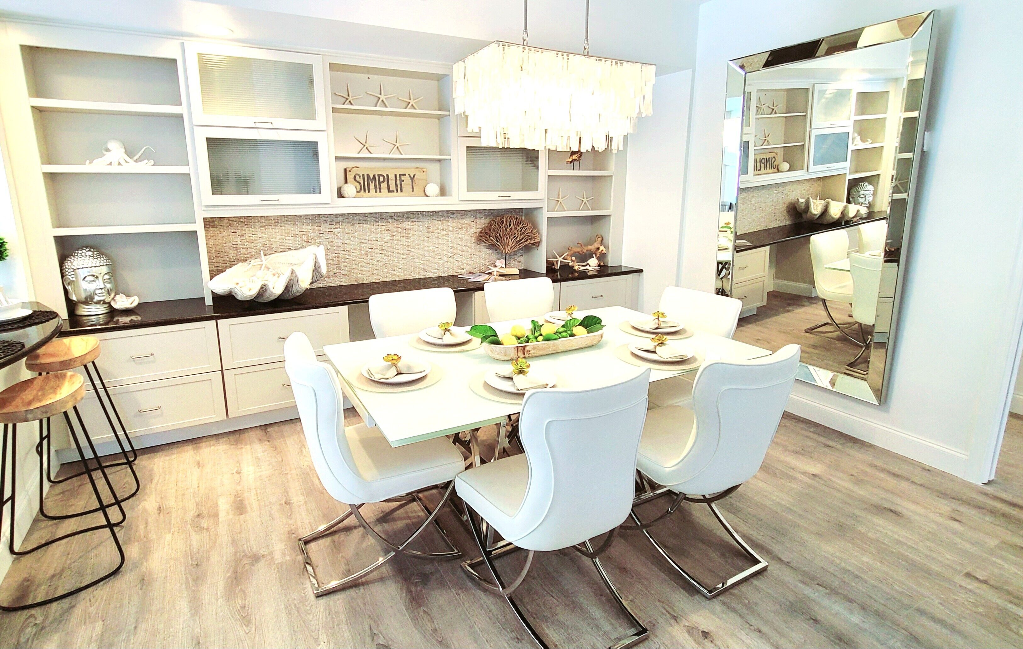 Un espace repas moderne avec une table blanche dressée pour quatre personnes, entourée de chaises blanches. On y trouve des étagères ornées d’objets décoratifs et un grand miroir au mur.