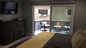 Un dormitorio con cama, cómoda, televisor y puerta corrediza de vidrio que da a un balcón con una silla y una mesita.