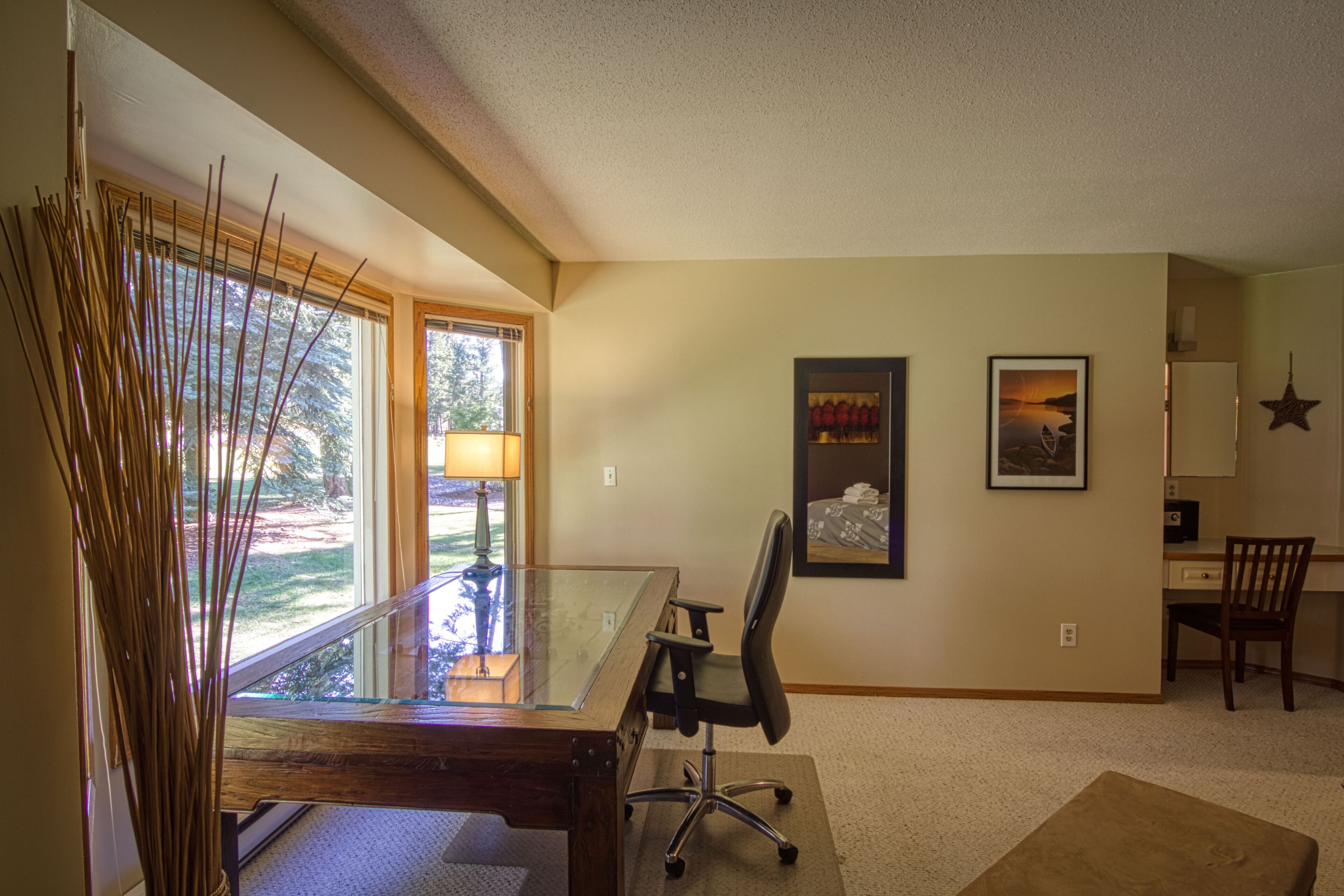 A spacious room with a glass desk, a comfortable office chair, a large window with a view of greenery, and a framed picture on the wall.