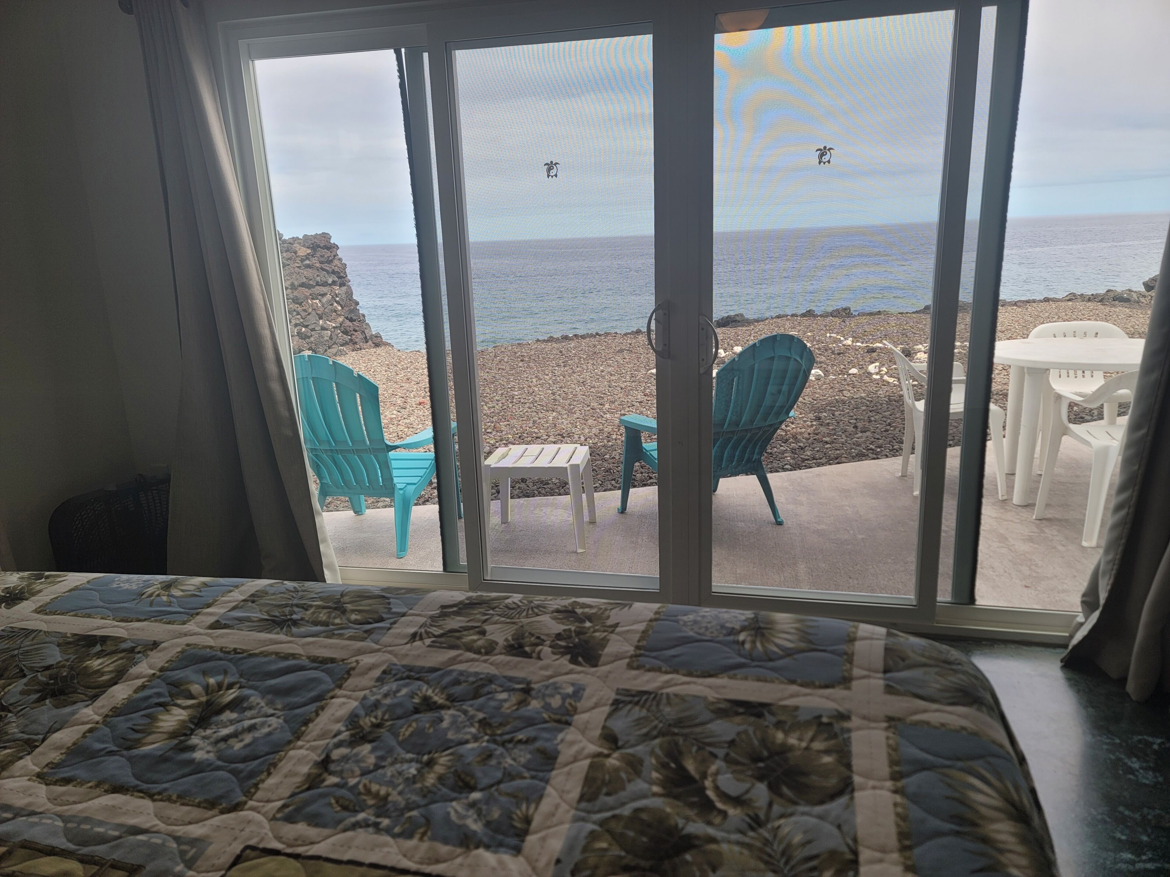A room with a bed, a view of a beach with chairs and a table outside.