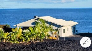 Una casa moderna con techo inclinado, ubicada sobre una superficie de roca volcánica oscura, con vista al mar tranquilo.