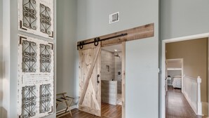 Un pasillo con una puerta de madera que da a un baño, piso de madera y una pared blanca con paneles metálicos decorativos.