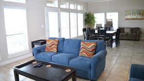A spacious living room with a blue sofa, a coffee table, and a dining area with chairs and a table. - Pirates Beach - Chez Antoine - Beautiful, Four Bedroom, Three Bath (Galveston)