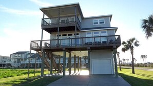 A multi-story residential building with a balcony on the upper floor, a garage at the base, and a grassy area in front. - Pirates Beach - Chez Antoine - Beautiful, Four Bedroom, Three Bath (Galveston)