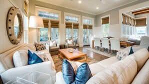 A spacious living room with a large sofa, a coffee table, and a dining area with a wooden table and chairs. - Coastal Chic Home with Exquisite Design, Outdoor Living, Golf Cart & Watercolor Beach Club Access (Santa Rosa Beach)