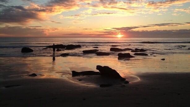 Una playa al atardecer con rocas y una persona de pie.
