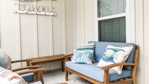 Ein gemütlicher Sitzbereich im Freien mit einem blauen Sofa, Holzstühlen und dekorativen Kissen. An der Wand sind ein schild in Fischform und ein Schild mit dem Schriftzug 'TOWELS' zu sehen.