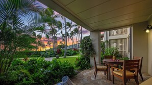 Une terrasse couverte avec un sol en pierre, une table en bois avec des chaises, un banc rembourré et une vue sur un jardin agrémenté de palmiers et d’un coucher de soleil.
